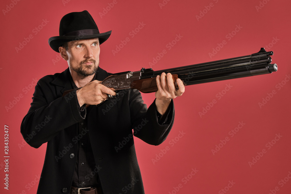 Middle-aged man with beard, mustache, in black jacket and hat, holding a gun while posing against a red background. Sincere emotions concept. Middle-aged man with beard, mustache, in black jacket and hat, holding a gun while posing against a red background. Sincere emotions concept.