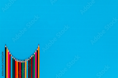 A bunch of crayons arranged like a parabola on a blue background , shot from above, aligned at the bottom left.