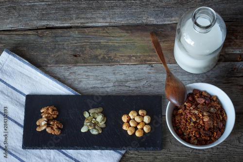 Crunchy honey granola bowl with flax seeds, cranberries, a bottle of milk and heaps of nuts on a black stand on a table, selective focus