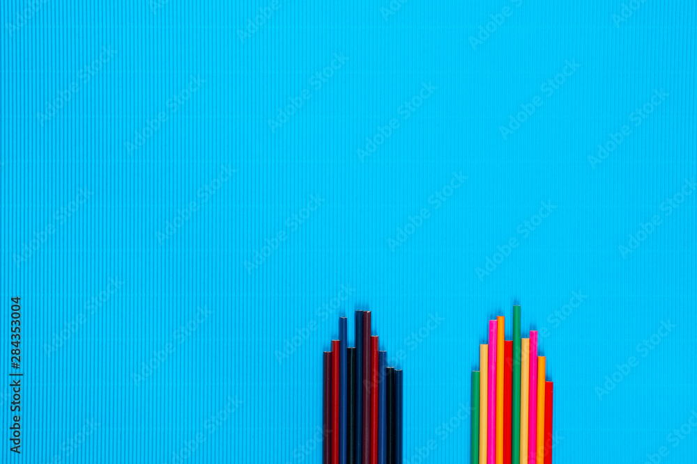 Two piles of crayons next to one another on a blue background, shot from above, arranged at the bottom left. Two piles of crayons next to one another on a blue background, shot from above, arranged at the bottom left.