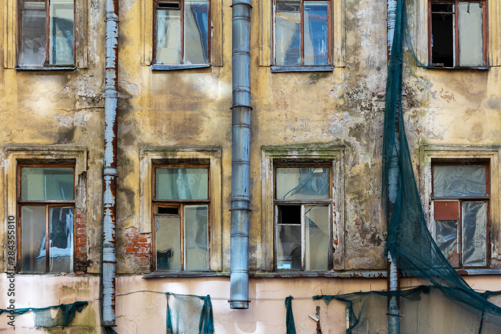 Naklejka premium An old residential building awaiting reconstruction or demolition. Facade with windows of crumbling plaster, scraps of green protective mesh and rusty drainpipes