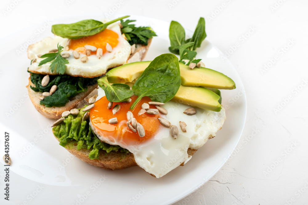 Fried eggs on a toasts bread with avocado, spinach and seeds on a white plate on the white background.