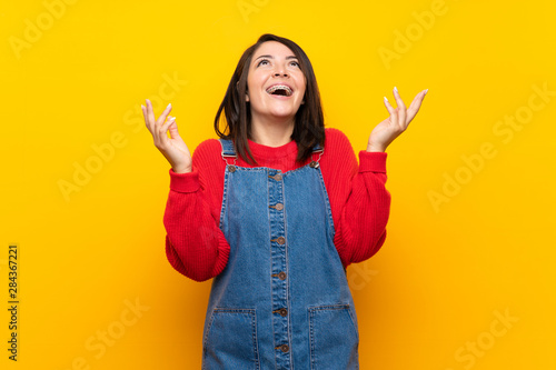 Young Mexican woman with overalls over yellow wall smiling a lot
