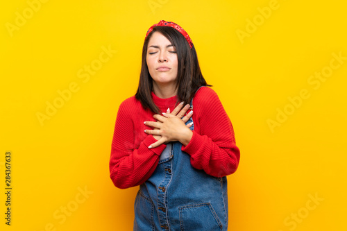 Young Mexican woman with overalls over yellow wall having a pain in the heart © luismolinero