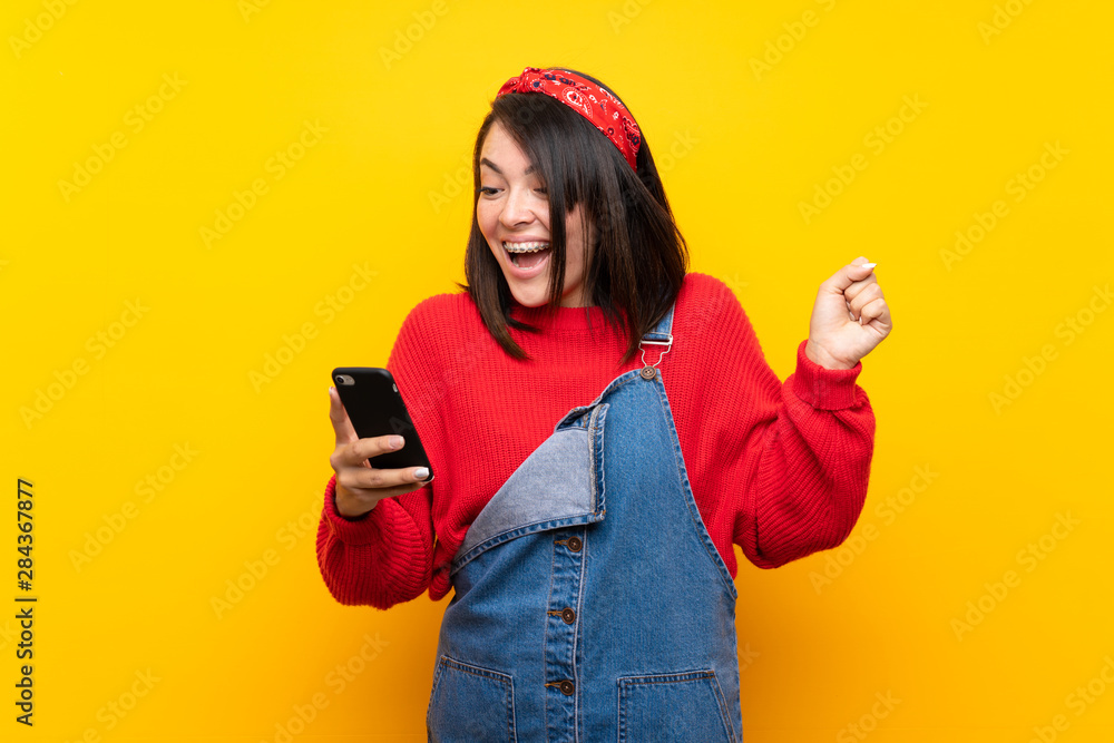 Young Mexican woman with overalls over yellow wall surprised and sending a message Young Mexican woman with overalls over yellow wall surprised and sending a message