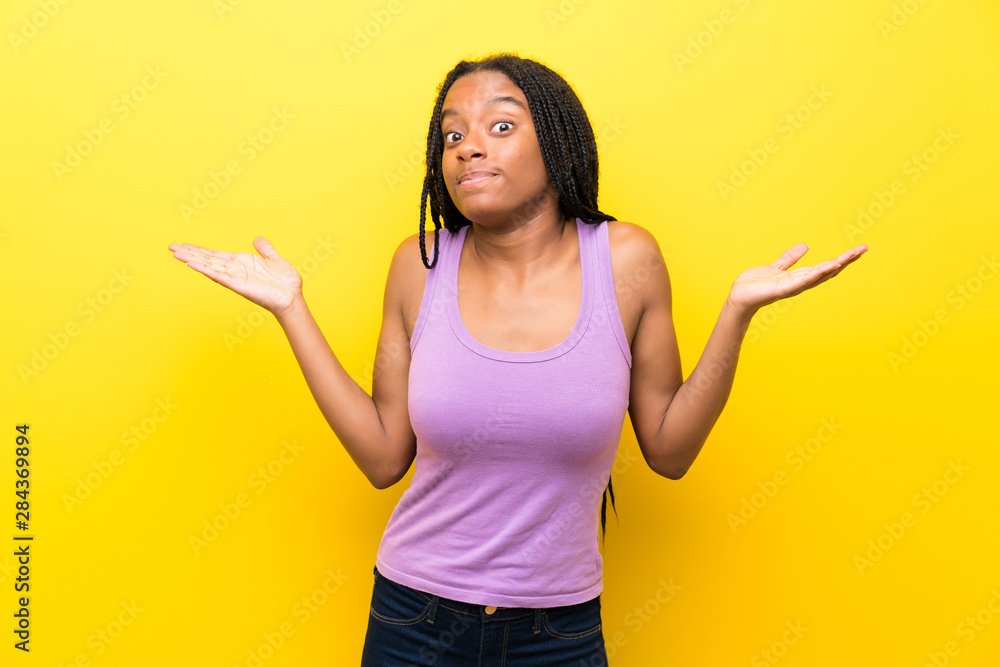 African American teenager girl with long braided hair over isolated yellow wall making doubts gesture African American teenager girl with long braided hair over isolated yellow wall making doubts gesture