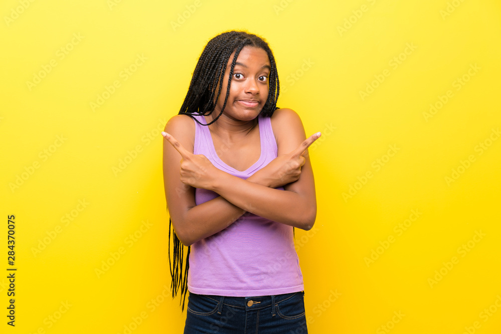 African American teenager girl with long braided hair over isolated yellow wall pointing to the laterals having doubts African American teenager girl with long braided hair over isolated yellow wall pointing to the laterals having doubts