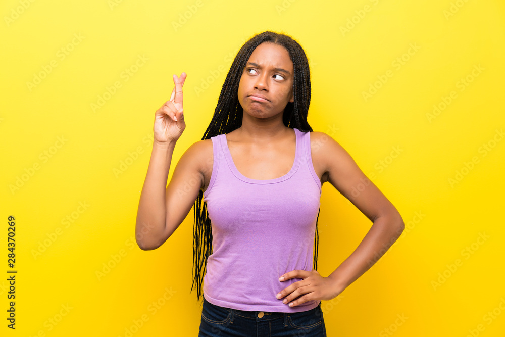 African American teenager girl with long braided hair over isolated yellow wall with fingers crossing and wishing the best African American teenager girl with long braided hair over isolated yellow wall with fingers crossing and wishing the best