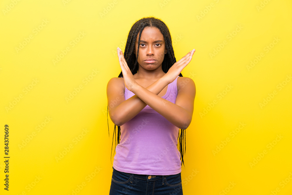 African American teenager girl with long braided hair over isolated yellow wall making NO gesture African American teenager girl with long braided hair over isolated yellow wall making NO gesture