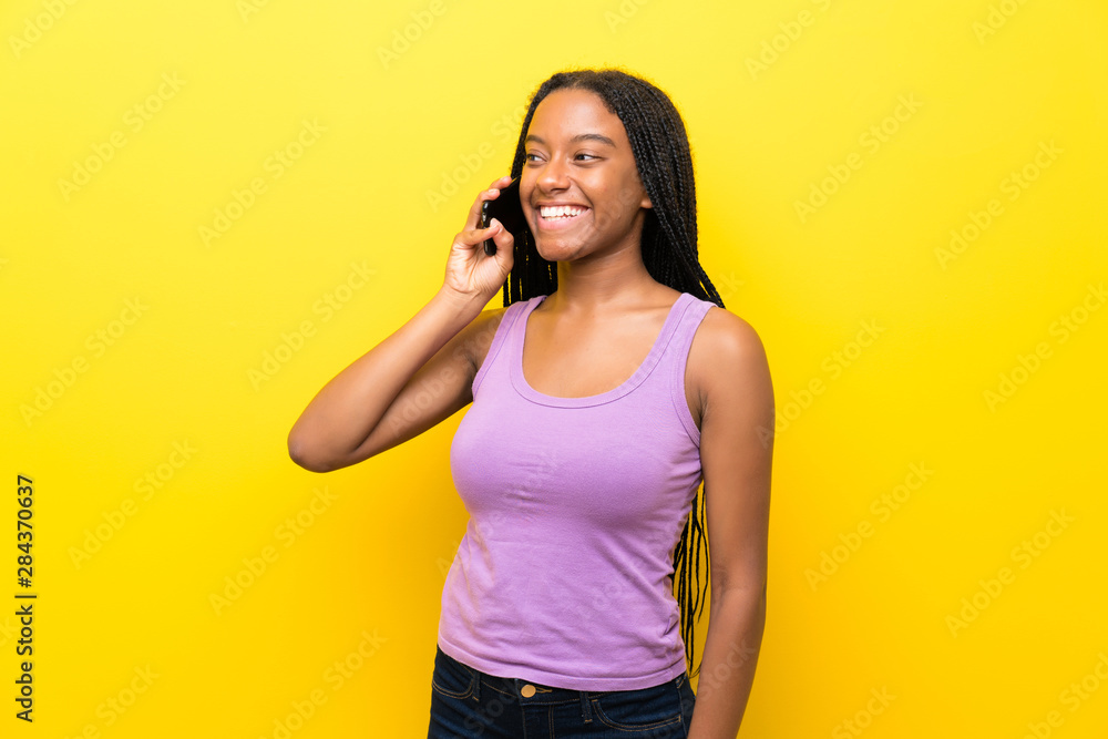African American teenager girl with long braided hair over isolated yellow wall keeping a conversation with the mobile phone African American teenager girl with long braided hair over isolated yellow wall keeping a conversation with the mobile phone