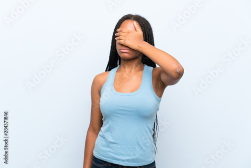 African American teenager girl with long braided hair over isolated blue background covering eyes by hands
