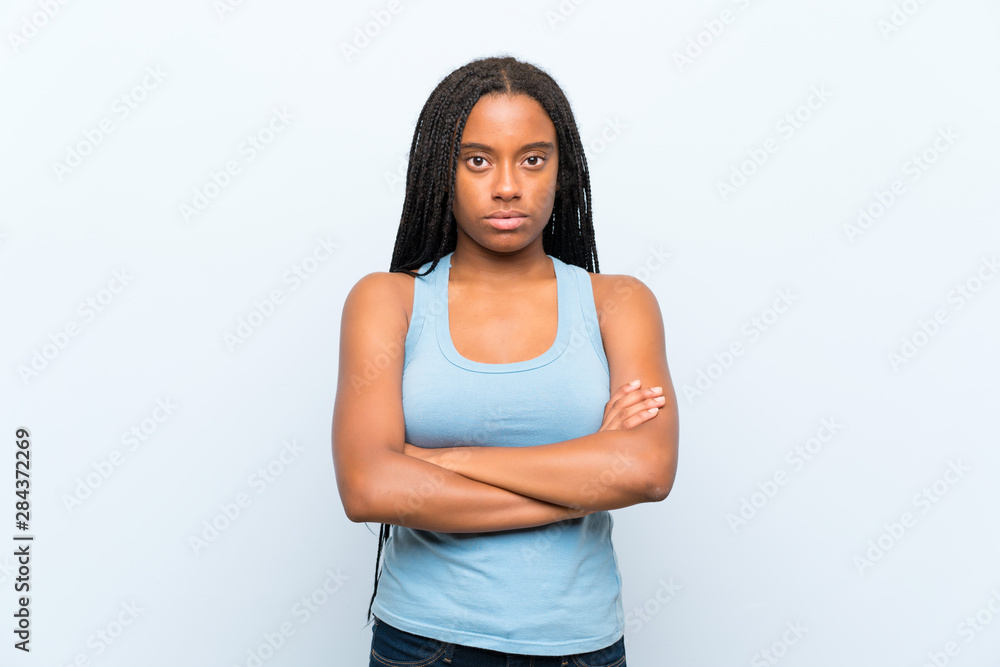 African American teenager girl with long braided hair over isolated blue background keeping arms crossed African American teenager girl with long braided hair over isolated blue background keeping arms crossed