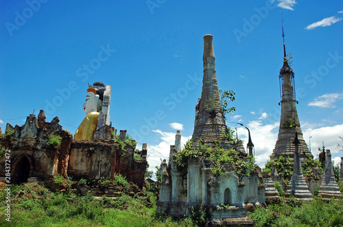 Myanmar, Inle, Ruins of an old buddhist temple and its sikhara tops; a smiling sitting buddha wearing a golden dress emerging from the rundown walls photo