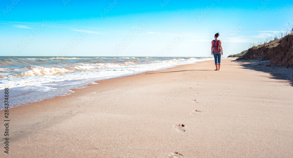young girl tourist with a red backpack on an empty sandy beach near the surf of the sea on a background of blue sky and clouds in summer