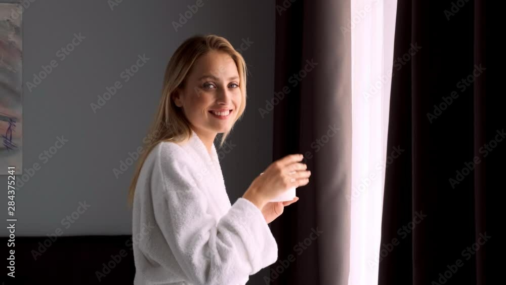 Medium shot of beautiful blond woman in bathrobe with cup of coffee happily looking in camera near window in hotel room Medium shot of beautiful blond woman in bathrobe with cup of coffee happily looking in camera near window in hotel room