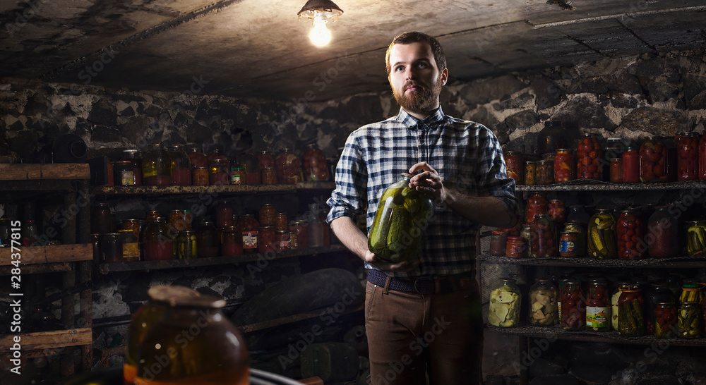 Bearded man in basement with jar of preservation cucumber in hands. Home Preservation concept. Bearded man in basement with jar of preservation cucumber in hands. Home Preservation concept.