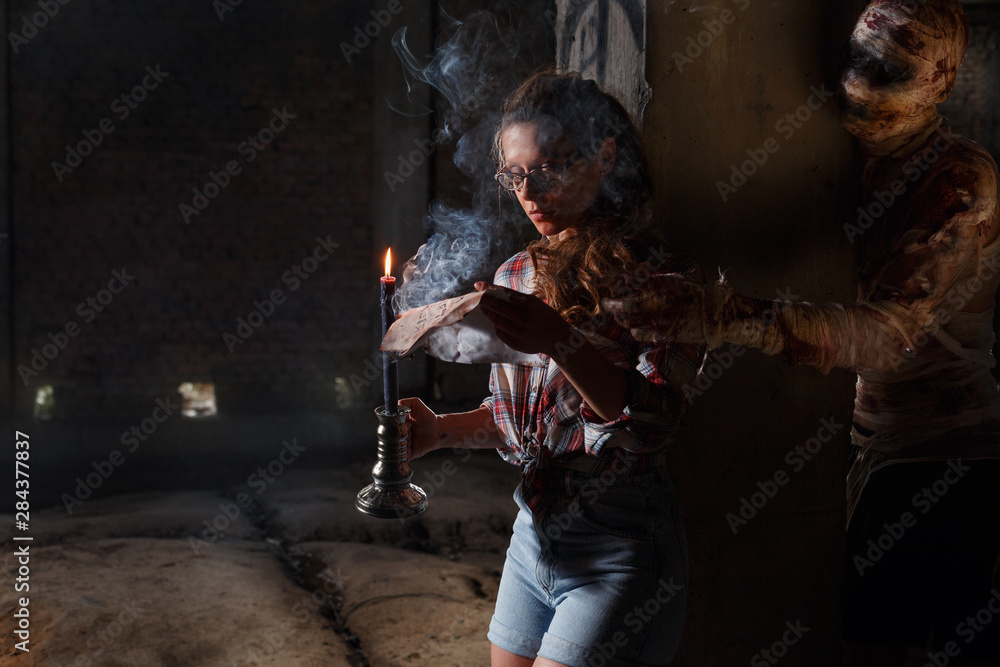 Naklejka premium Adventure seeker looking into the ancient map of treasure while alive mummy sneaked up behind. Horror concept