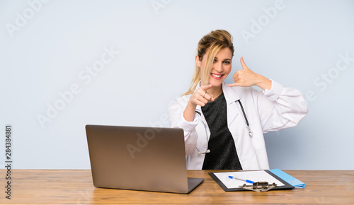 Blonde doctor woman making phone gesture and pointing front