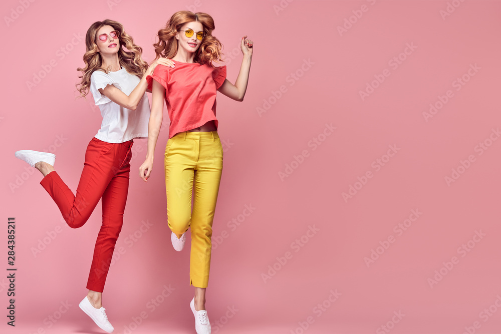 Fototapeta premium Fashion. Two Inspired woman laughing dance. Shapely joyful friend Having Fun, Stylish fashionable summer outfit. Carefree Girl friends with curly hair jump dancing in Studio, happy positive emotion
