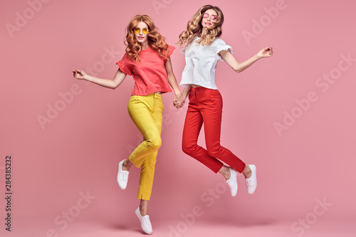 Fashion. Two Inspired woman laughing dance. Shapely joyful friend Having Fun, Stylish fashionable summer outfit. Carefree Girl friends with curly hair jump dancing in Studio, happy positive emotion