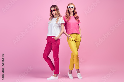 Fashion. Two Beautiful woman in Trendy summer outfit posing on pink. Graceful sisters friends in yellow coral pants with curly hair  make up. Colorful creative fashionable Indoor shot