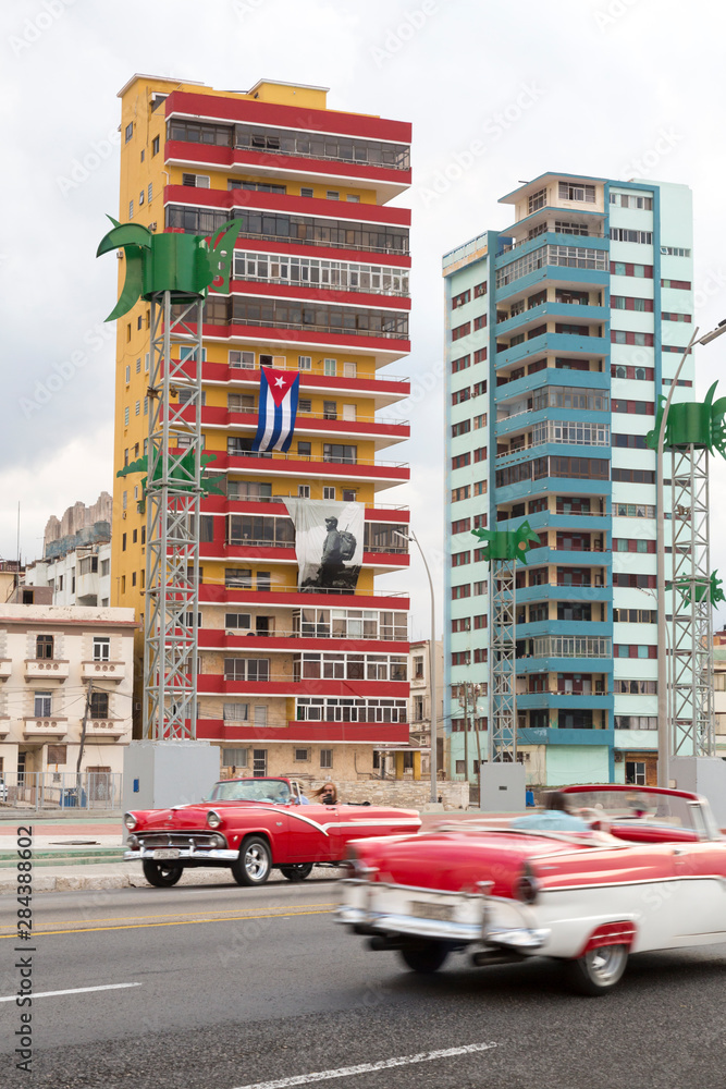Cuba, Havana. Classic cars pass apartment buildings.