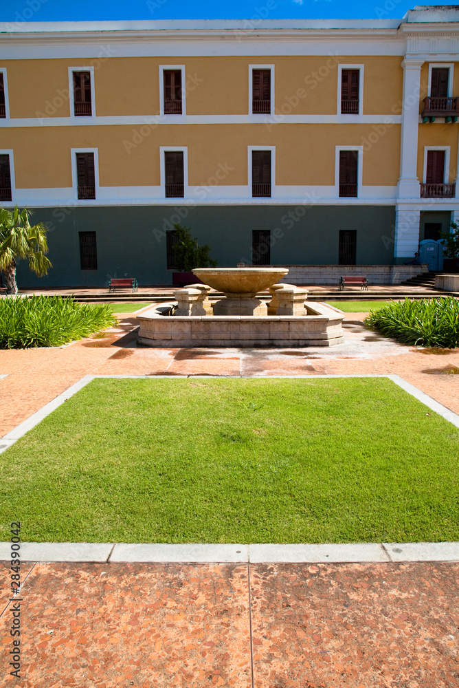 Obraz premium San Juan, Puerto Rico - A hotel fountain in a courtyard is sitting with no water in it. A patch of grass can be seen in the foreground bordered by walkways.