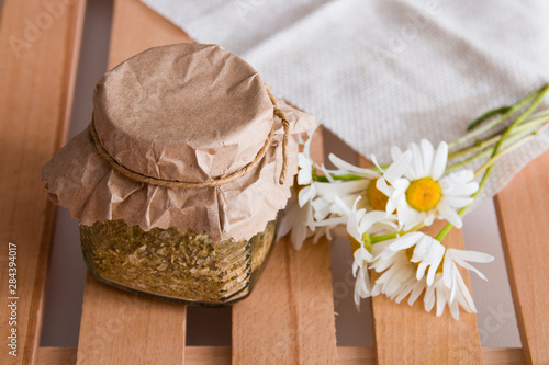 a jar of herbal tea stands on a wooden tray of rekk with a daisy flower and a beige coarse cloth copy space top view
