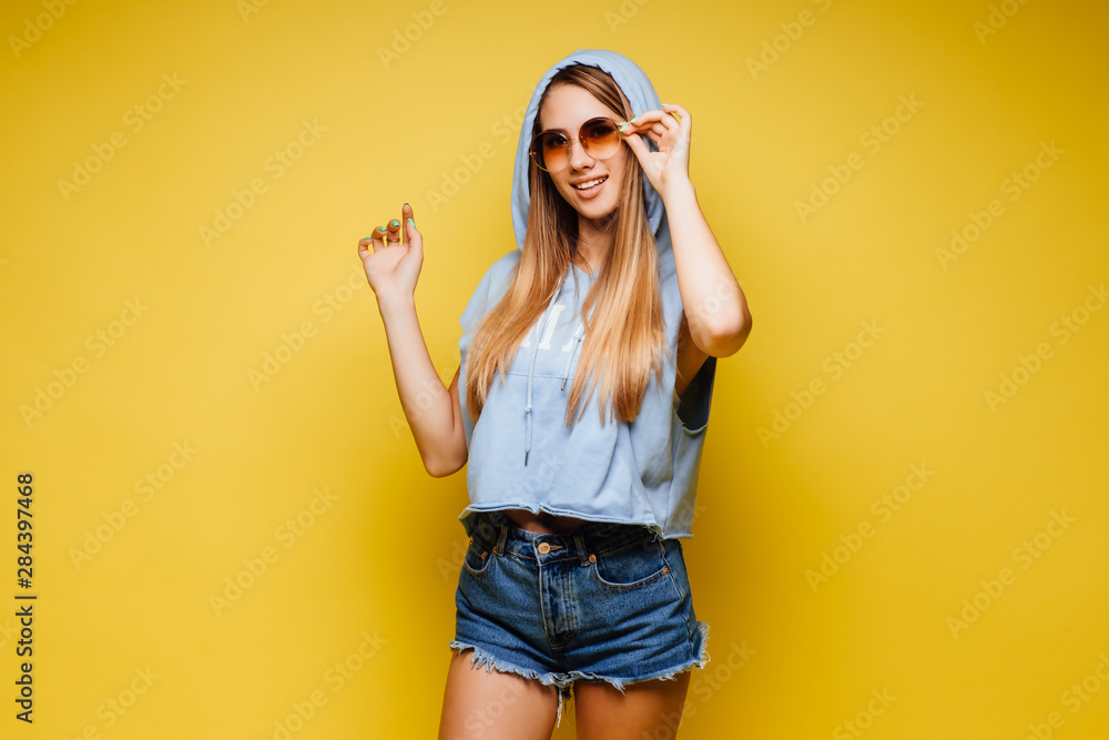 Young blonde,cute woman over isolated yellow wall wearing in sportive style in sunglasses posing. Young blonde,cute woman over isolated yellow wall wearing in sportive style in sunglasses posing.
