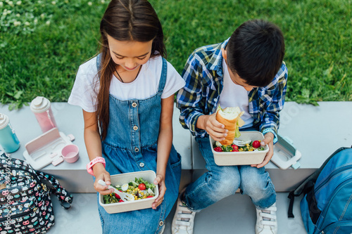 Little brother and sister siting on the grass together in summer park with lunch boxes and smart watch on hands.