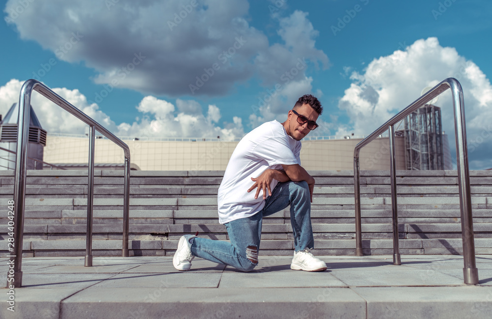 guy dancer in a white T-shirt, jeans, dancing break dance in summer in the city, in sunglasses, white sneakers background building clouds guy dancer in a white T-shirt, jeans, dancing break dance in summer in the city, in sunglasses, white sneakers background building clouds
