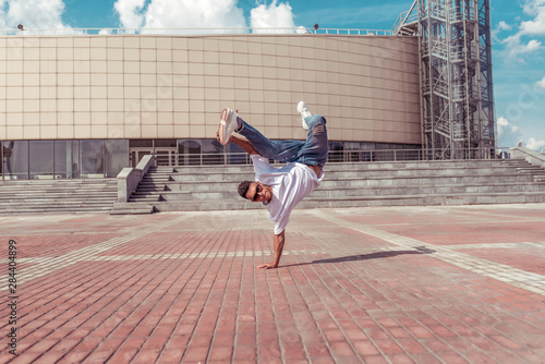 young guy dancer in white T-shirt, jeans, stands on one arm, in summer city, sunglasses, background building, active hip hop, youth lifestyle, free space for text, acrobatic stunt brake dance