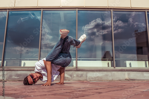 athlete guy dancer in a white T-shirt, jeans, on his head, summer in city, in sunglasses, background glass windows, active hip hop, youth lifestyle, free space for text, acrobatic trick break dance