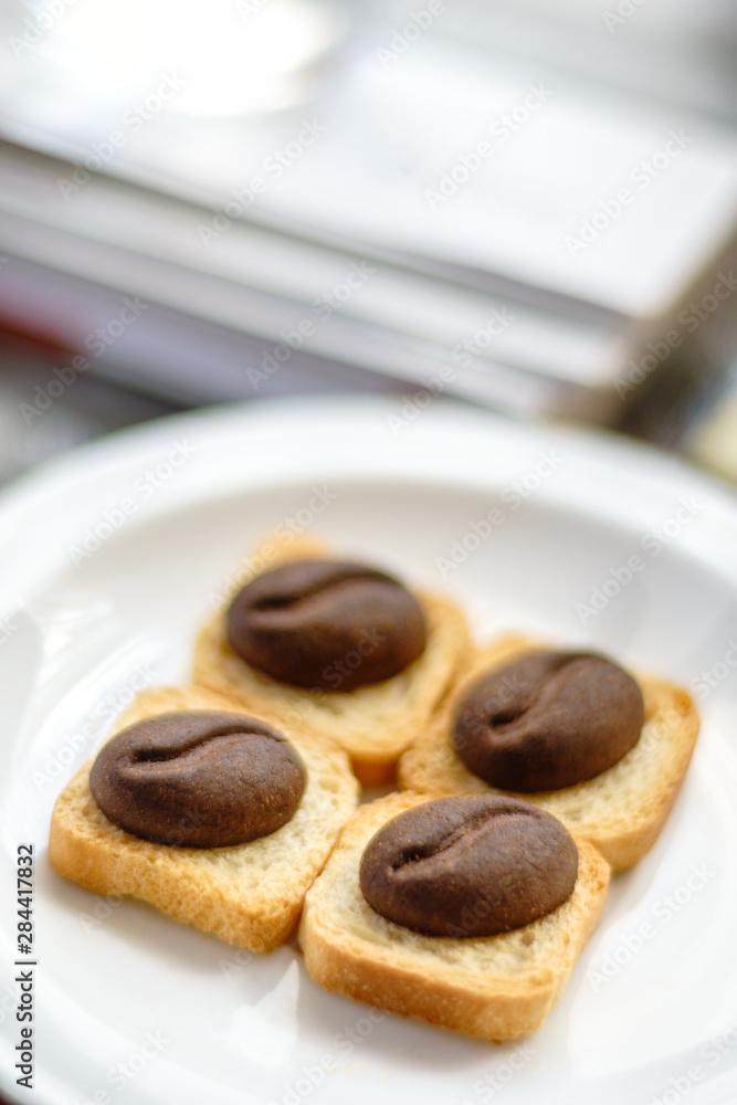 Four brown cookies with shape of cocoa beans lay on square breadcrumbs in white plate. Four brown cookies with shape of cocoa beans lay on square breadcrumbs in white plate.