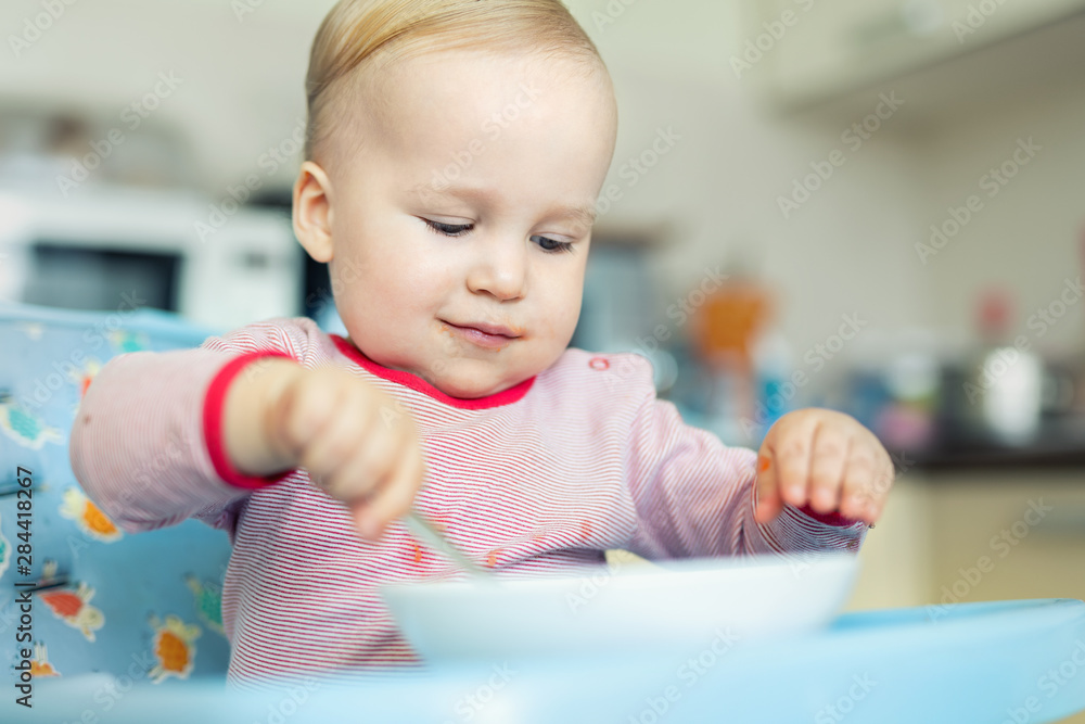 Adorable cute caucasian blond toddler boy eating tasty vegetable soup in chair at kithcen indoor. Mother feeding baby healthy food with spoon. Natural children nutriotion and diet