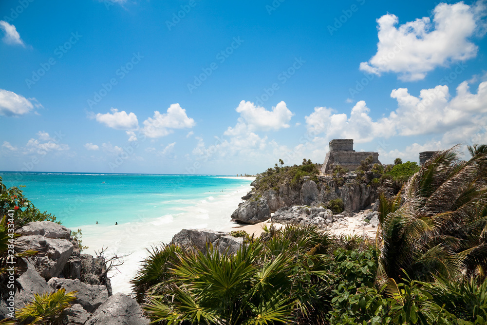 Cancun, Quintana Roo, Mexico - A white sand beach behind tropical foliage. An old stone structure can be seen on a hill in the distance.