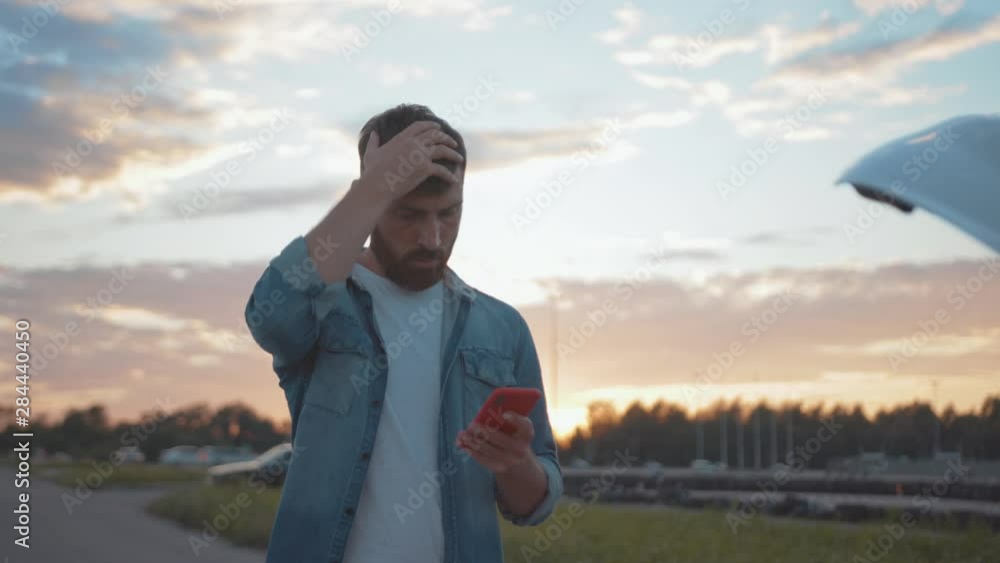 Young man driver is upset with car trouble calling services for assistance standing looking on breakdown vehicle standing on roadside.