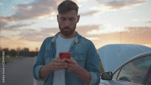 Nervous young man using smartphone calling car assistance services standing by breakdown automobile on the roadside at sunset.