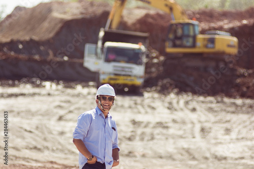 portrait mining supervisor or engineer in hardhat posing with hand inside pocket in front of white truck at coal and soil mining site in daylight