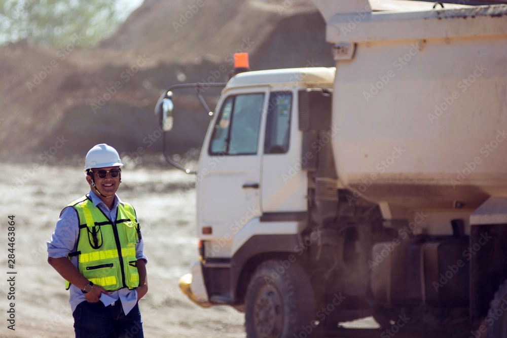 portrait mining worker or engineer in reflective vest and hardhat posing with hand inside pocket in front of white truck at coal and soil mining site