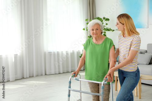 Caretaker helping elderly woman with walking frame indoors photo