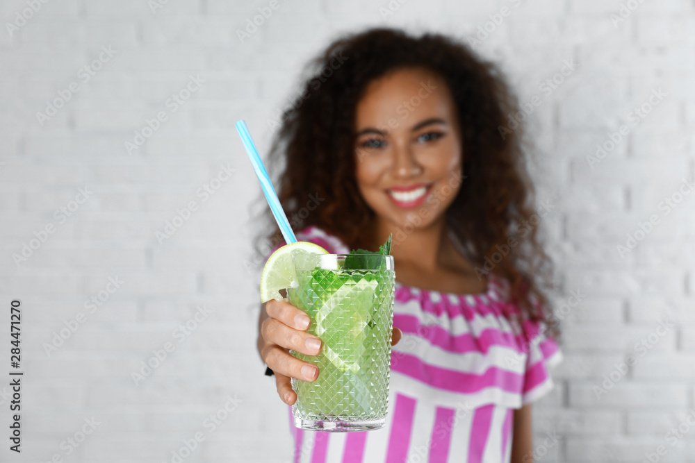 Happy African-American woman with glass of natural lemonade near white brick wall, closeup. Detox drink Happy African-American woman with glass of natural lemonade near white brick wall, closeup. Detox drink