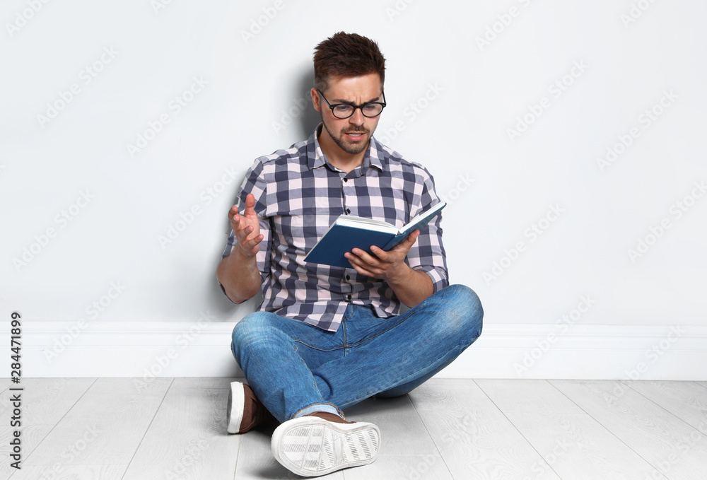 Handsome young man reading book on wooden floor near light wall