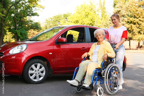 Senior woman in wheelchair with granddaughter near car outdoors
