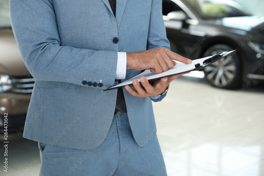 Young salesman with clipboard in modern car dealership, closeup