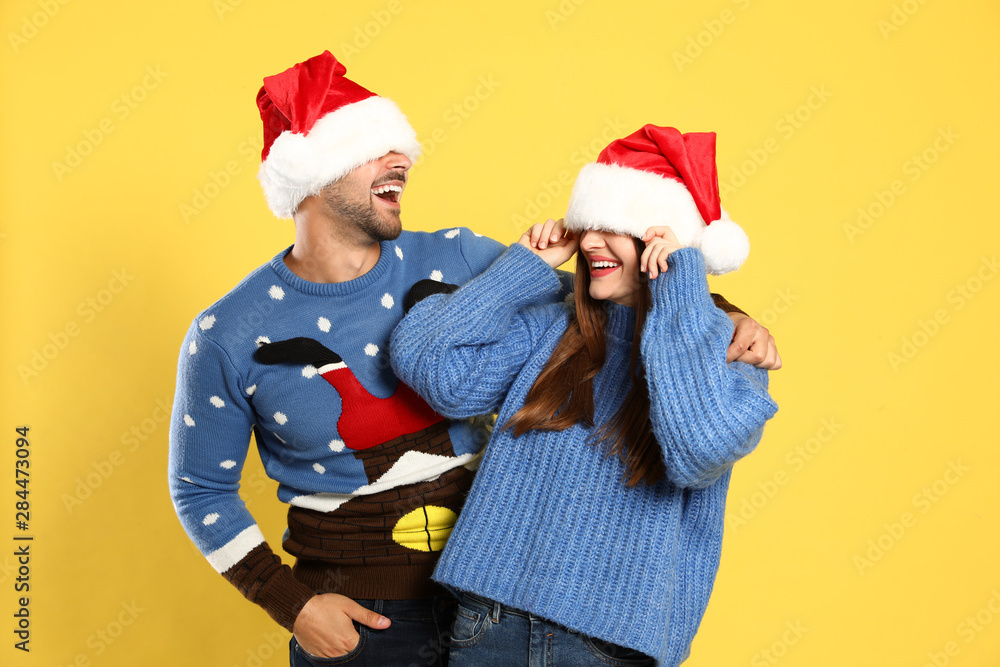 Fototapeta premium Couple wearing Christmas sweaters and Santa hats on yellow background