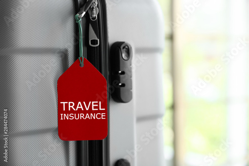 Stylish suitcase with travel insurance label on blurred background, closeup. Space for text