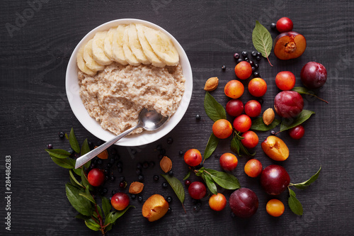 Oatmeal with banana in white deep bowl on dark wooden table. Healthy breakfast with oatmeal and fresh organic berries, table decoreted with plums, aliches and green leaves. Healthy food concept. photo