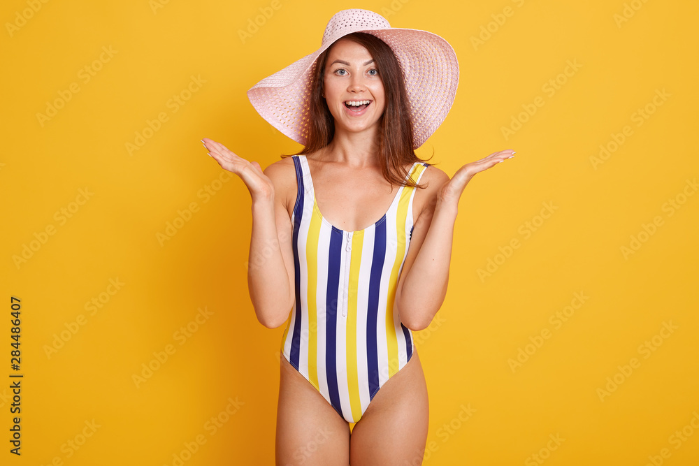 Close up portrait of surprised woman in summer hat and striped swimming suit, staring with bugged eyes and opens mouth from surprisement, standing against yellow background. People emotions concept. Close up portrait of surprised woman in summer hat and striped swimming suit, staring with bugged eyes and opens mouth from surprisement, standing against yellow background. People emotions concept.