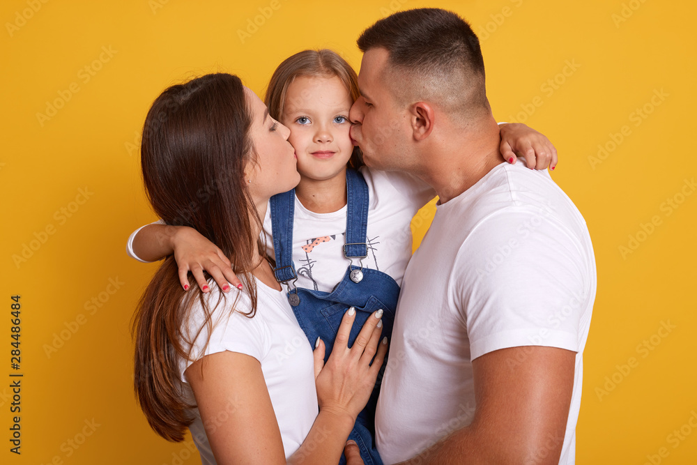 Fototapeta premium Happy young family kissing ther little charming daughter. Cute child girl sitting close to parents and looks at camera. Mother, father and kid expressing happyness, glad to be together. Family concept